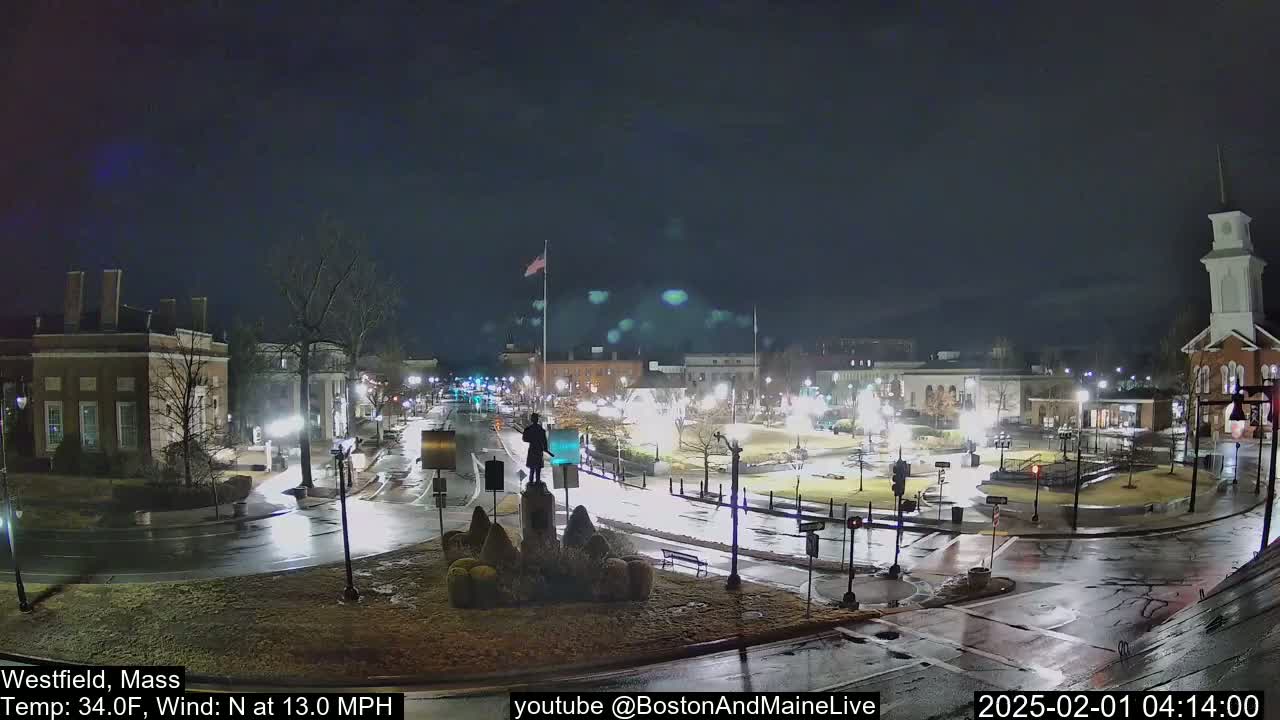 A nighttime view of a town square with a statue in the center, wet roads, and a few cars, under a dark, overcast sky.