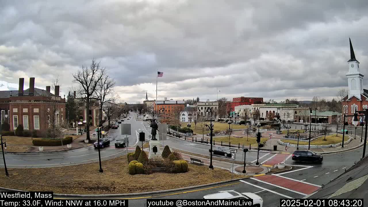A town square with several brick buildings, a monument, and sparse traffic under a cloudy sky.