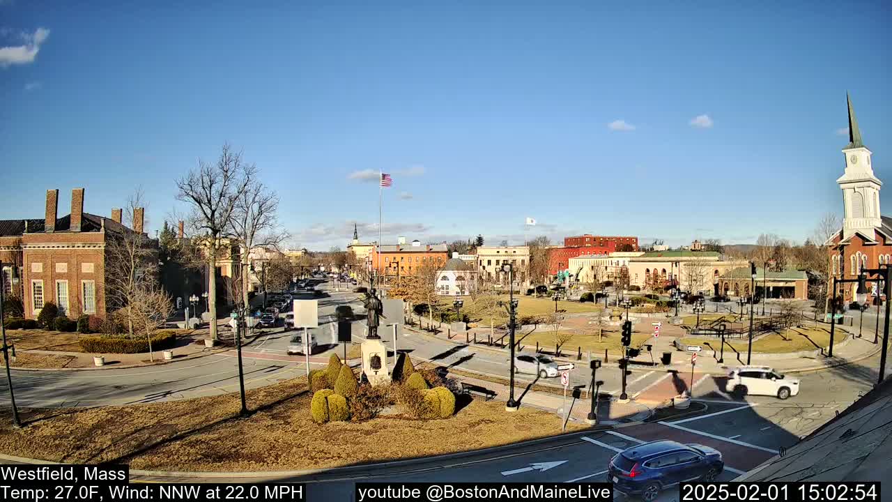 A sunny town square with brick buildings, a statue, and sparse traffic under a clear blue sky.
