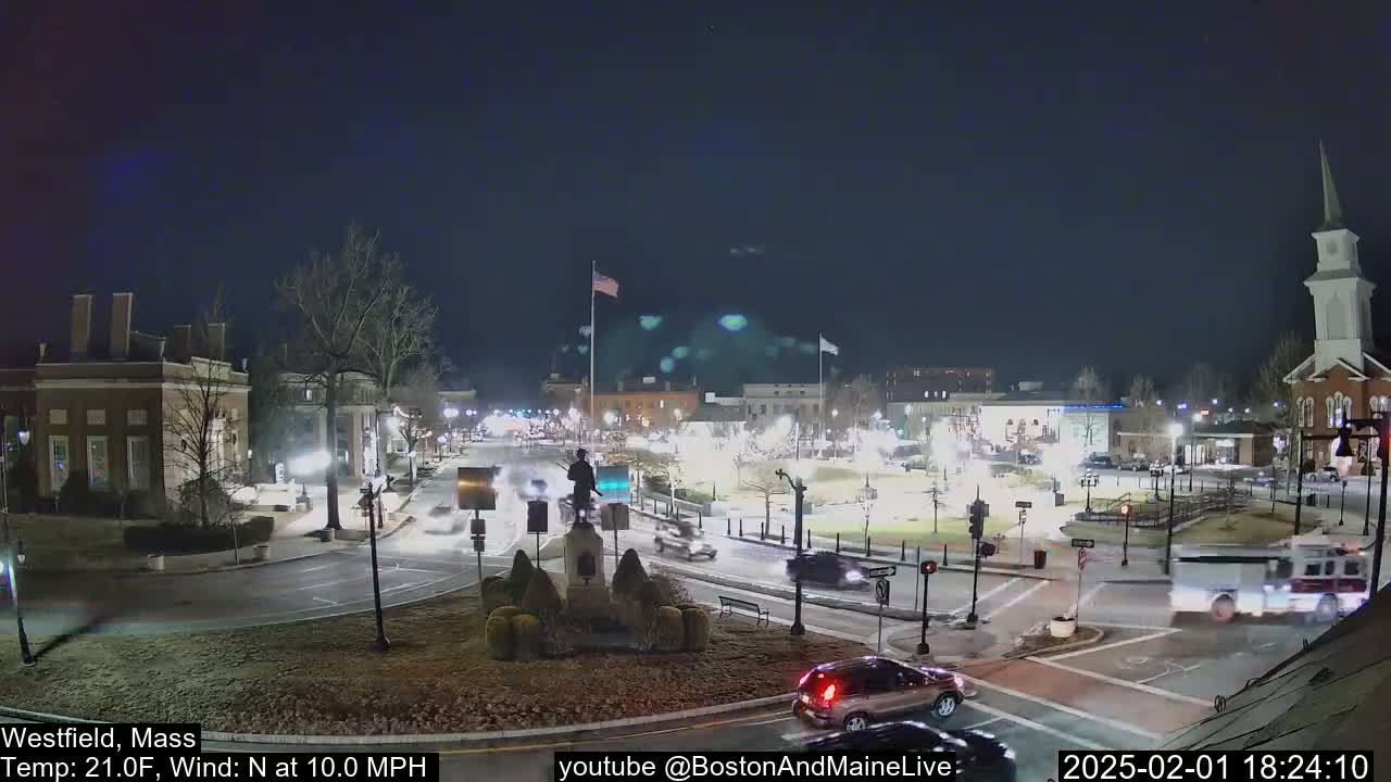 A nighttime view of a town square with a statue in the center, cars driving around the square, and buildings lit up under a clear, dark sky with a north wind of 10 mph and a temperature of 21°F.