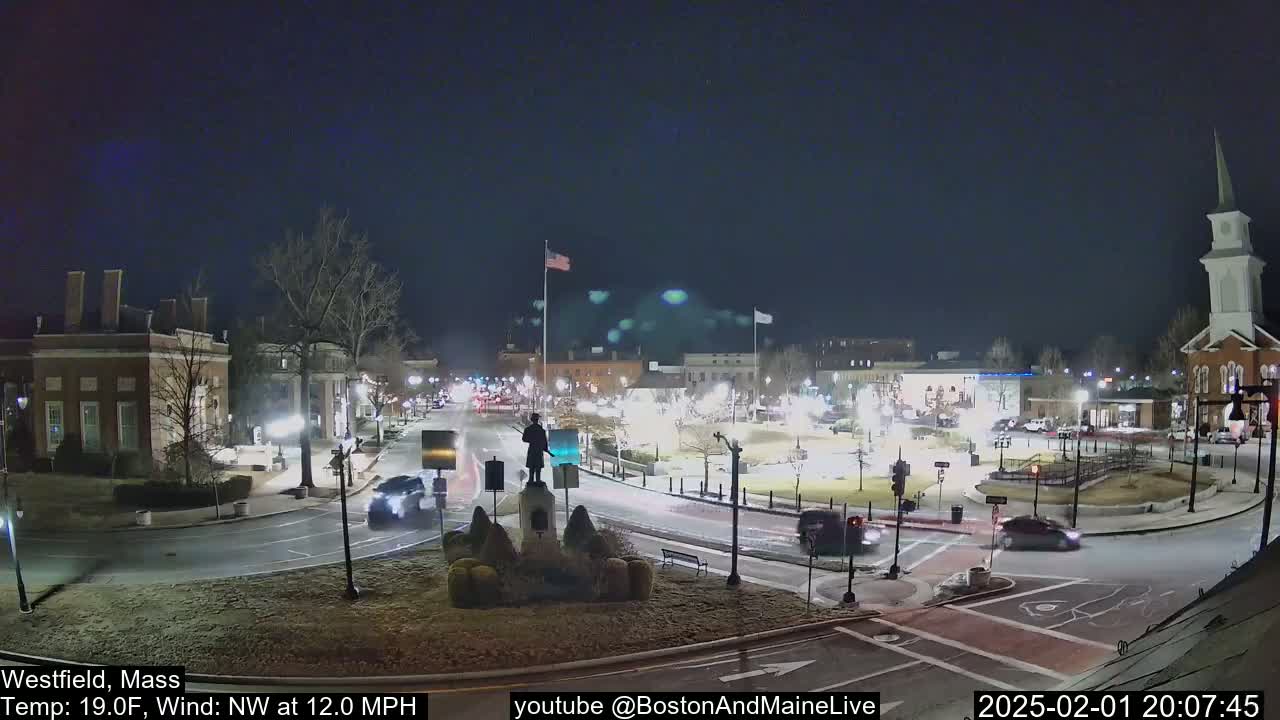 A nighttime view of a town square with a statue at its center, surrounded by buildings and roads with cars, under a clear, dark sky with a temperature of 19°F and a northwest wind of 12 mph.