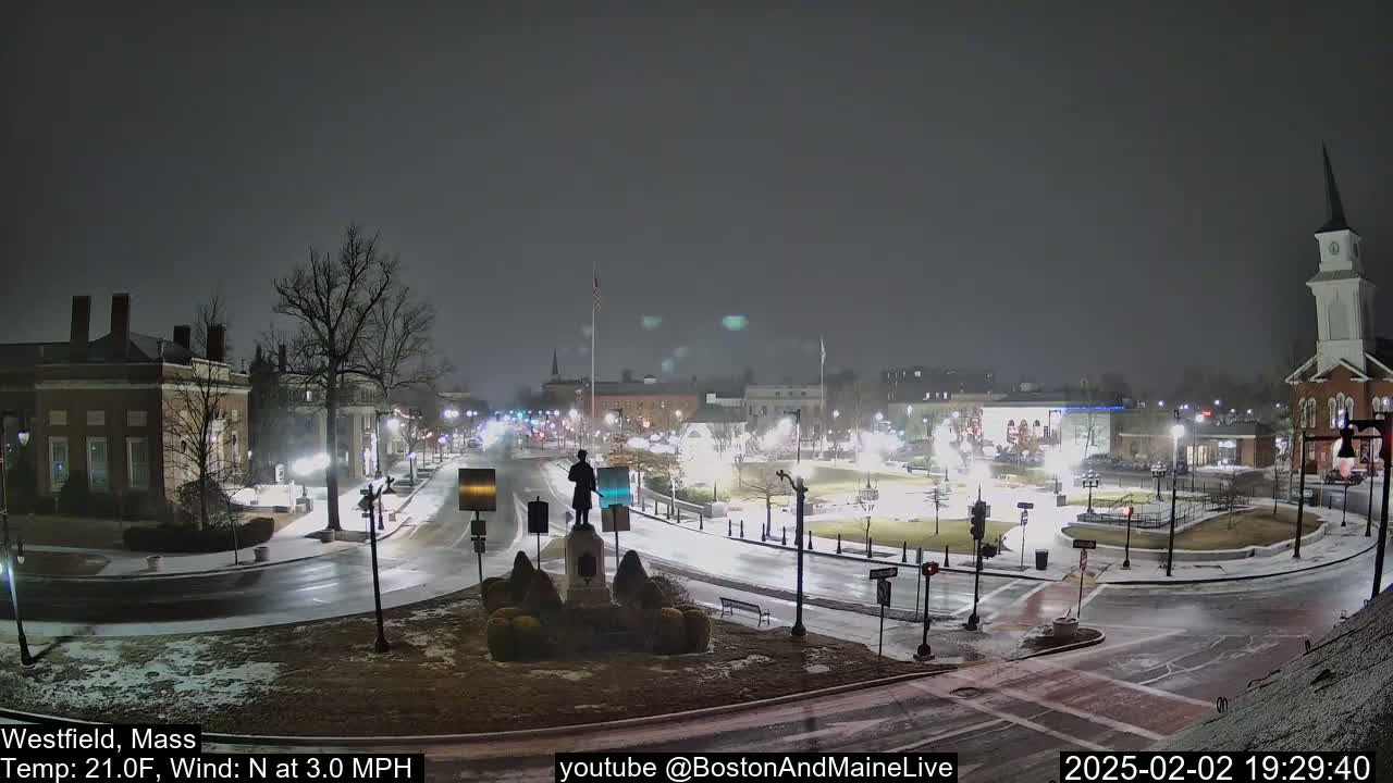 A snow-covered town square at night, illuminated by streetlights, features a statue in the center and a church in the background.