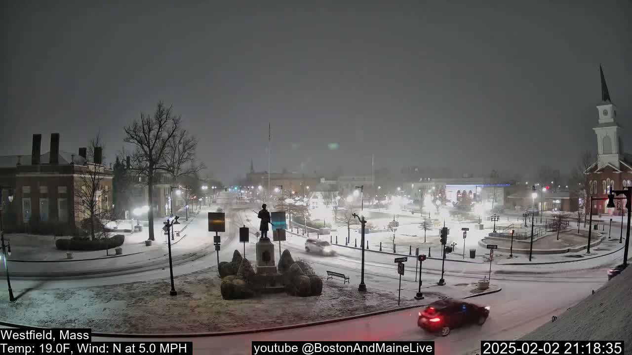 A snow-covered town square at night, with cars driving through the falling snow and a statue in the center.