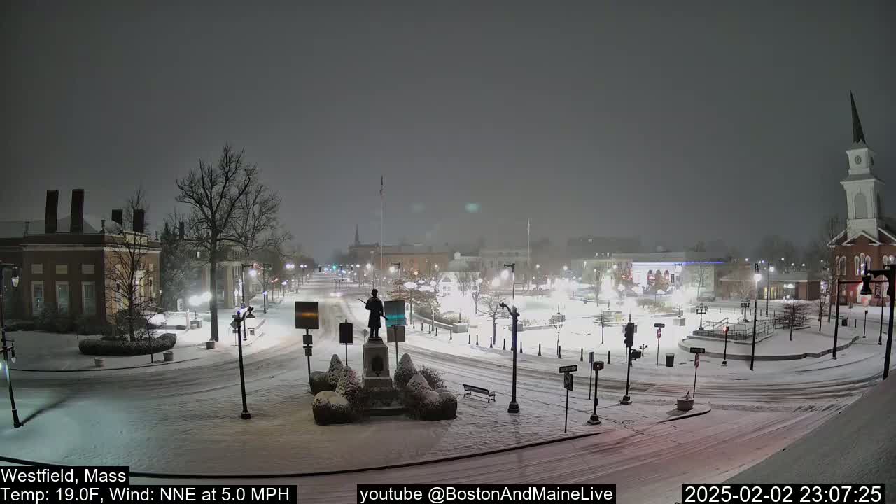 A snow-covered town square at night, featuring a statue, buildings, and a church steeple under a lightly falling snow.