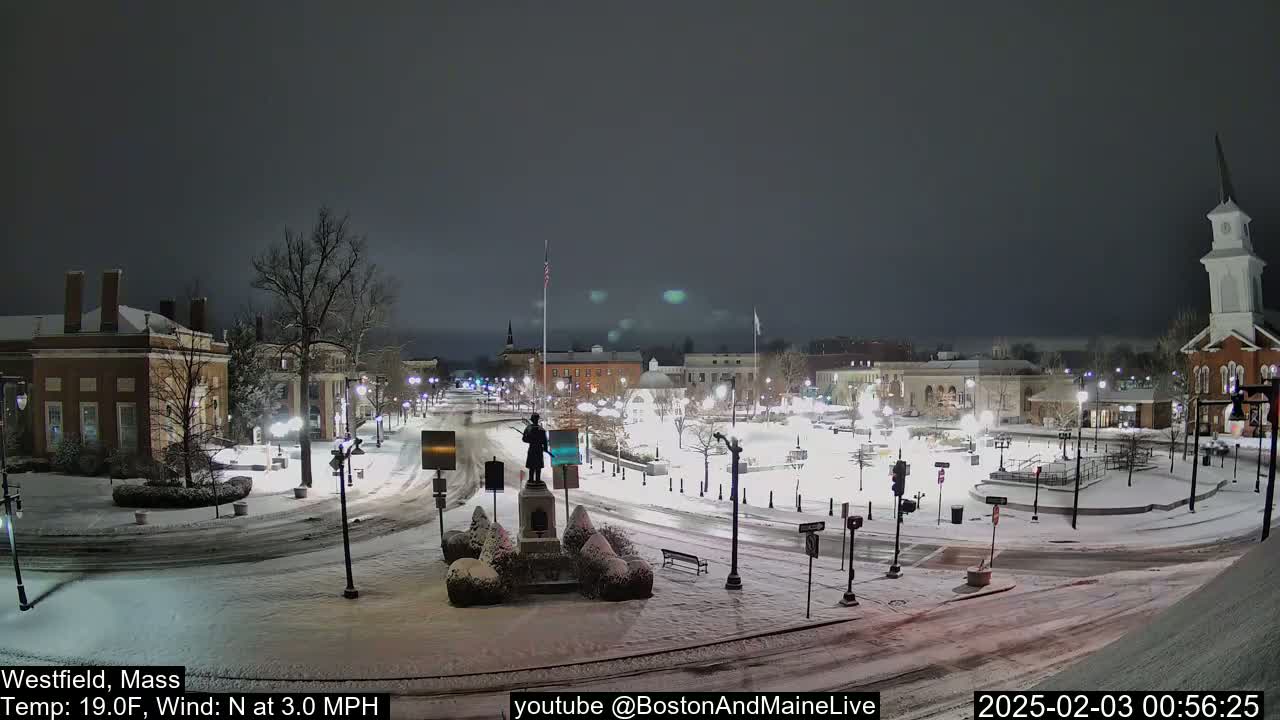 A snow-covered town square at night, illuminated by streetlights, features a statue in the center and several buildings surrounding it.