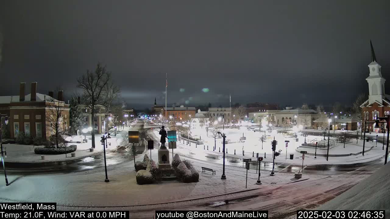A snow-covered town square at night, illuminated by streetlights, features a statue in the center and calm weather conditions.