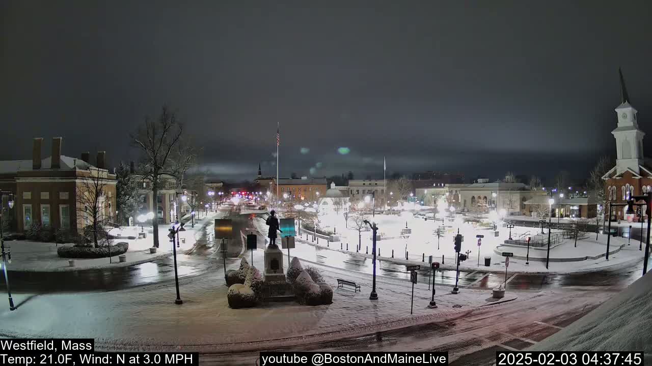 A snow-covered town square at night, illuminated by streetlights, with a statue in the center and buildings surrounding it under a dark, overcast sky.
