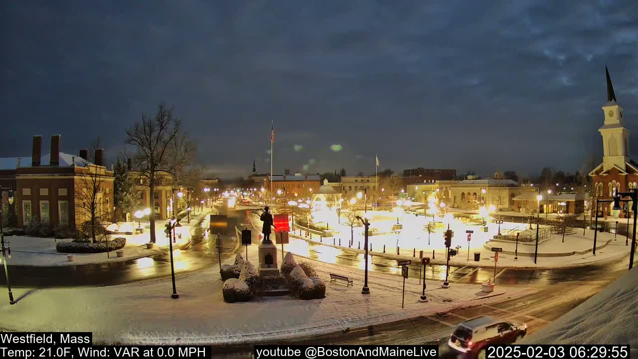 A snow-covered town square at night, illuminated by streetlights, with cars driving on wet roads.
