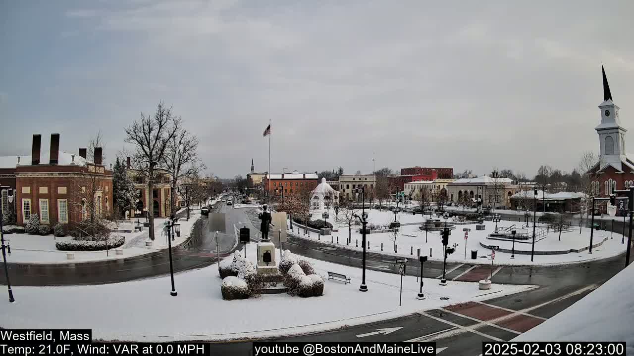 A snow-covered town square with brick buildings, a statue, and a church steeple under an overcast sky.