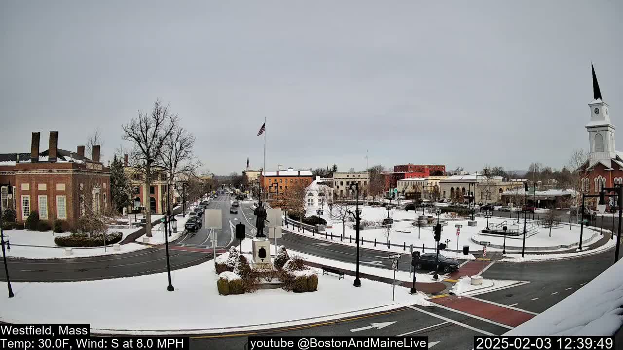 A snow-covered town square with brick buildings, a statue, and a church steeple, under a cloudy sky.