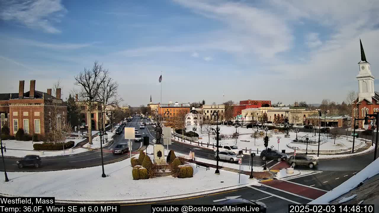 A snow-covered town square with brick buildings, a church steeple, and cars driving on roads under a partly sunny sky.