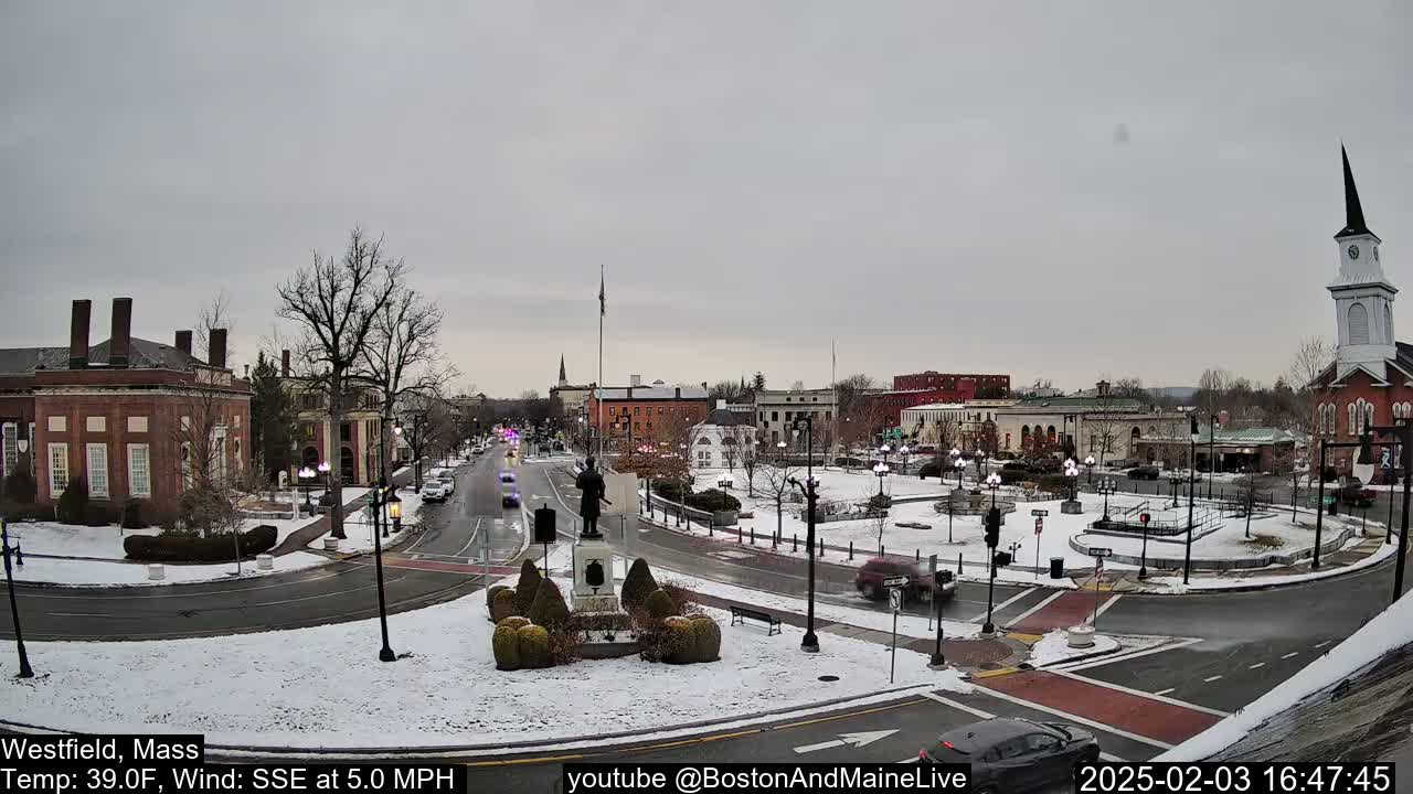 A snow-covered town square with a statue in the center, surrounded by brick buildings and cars, under an overcast sky.
