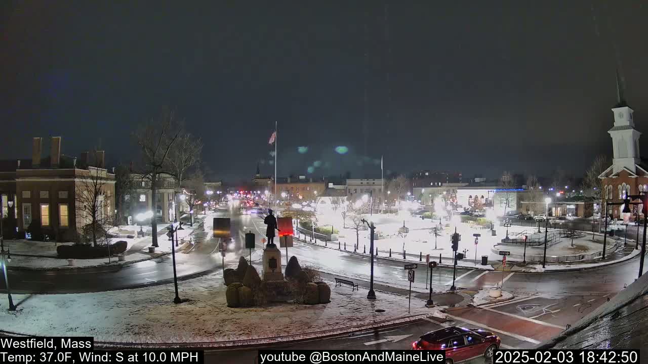 A nighttime view of a town square with a statue in the center, light snow on the ground, and cars driving on wet roads under a dark sky.