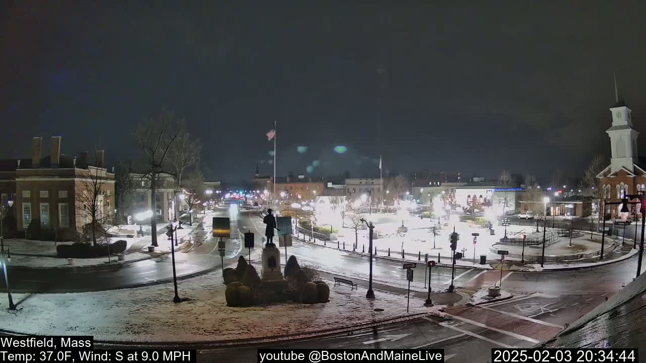 A snow-covered town square at night, illuminated by streetlights, with a statue in the center and surrounding buildings.