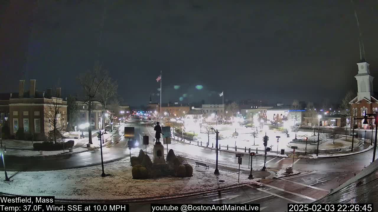 A nighttime view of a snow-covered town square with a statue at its center, under a dark sky with a SSE wind at 10 mph and a temperature of 37°F.