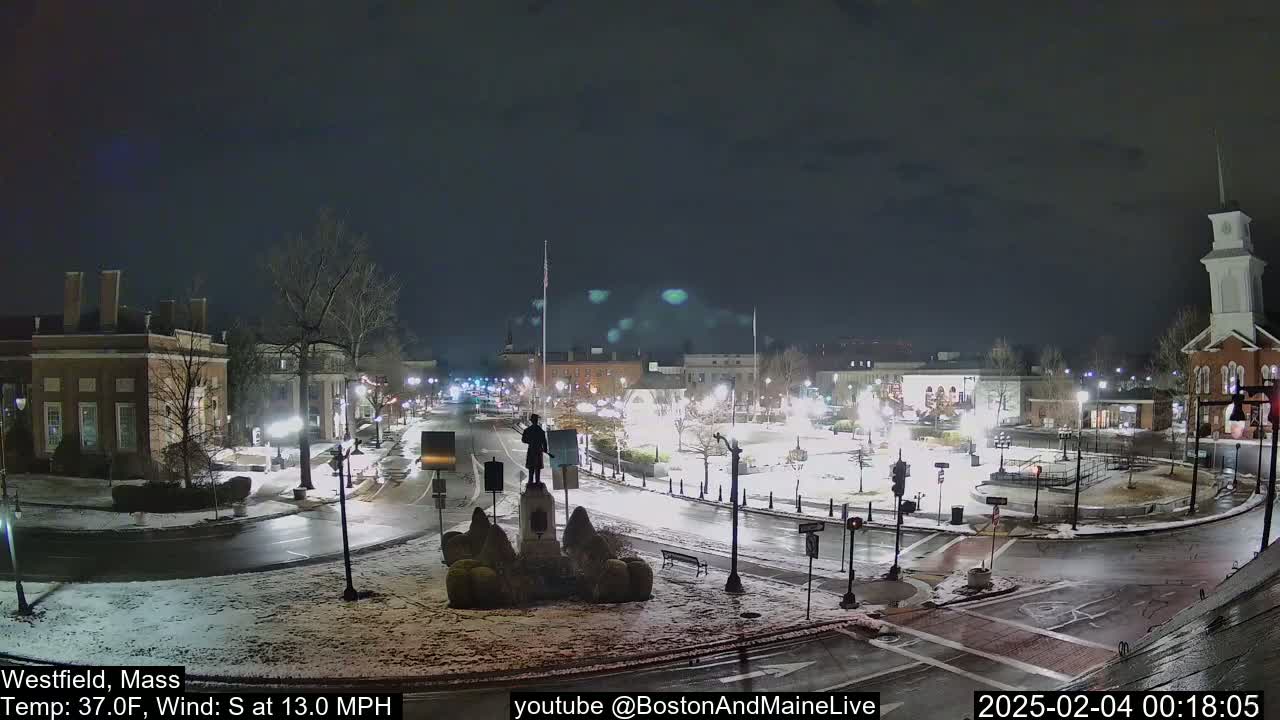 A snow-covered town square at night, with a statue in the center, is illuminated by streetlights under a dark sky.