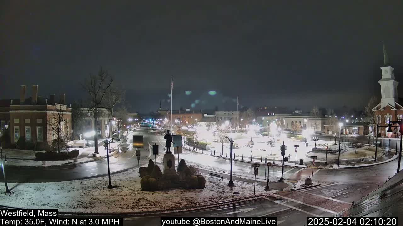 A snow-covered town square at night, illuminated by streetlights, with a statue in the center and buildings surrounding it.