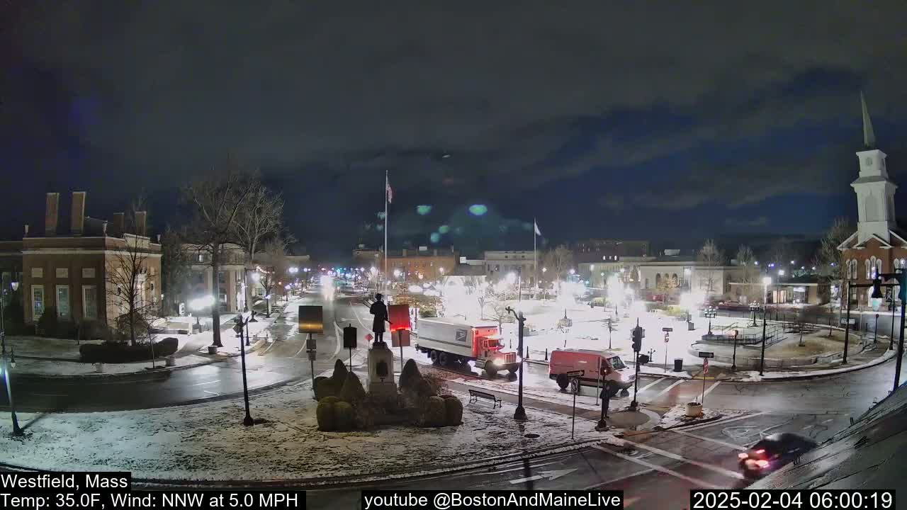 A snow-dusted town square at night, with vehicles at an intersection, under a partly cloudy sky.