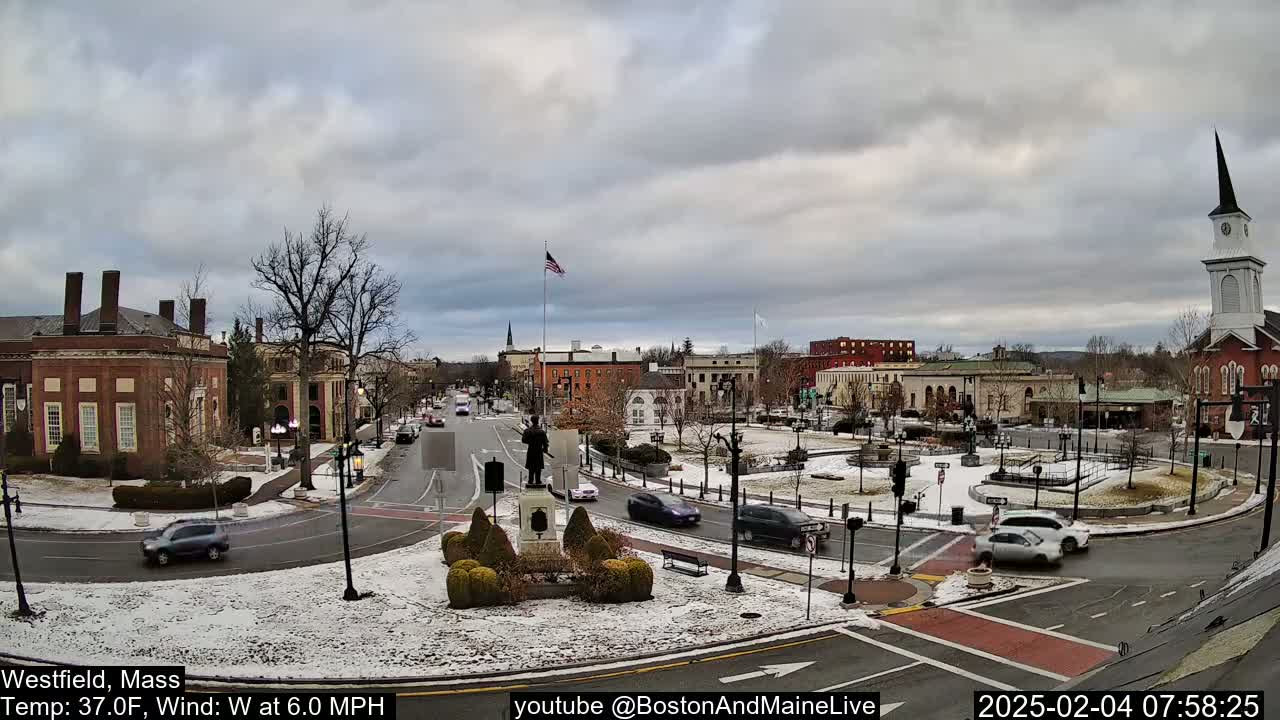 A snow-dusted town square with brick buildings, a statue, and several cars under an overcast sky.