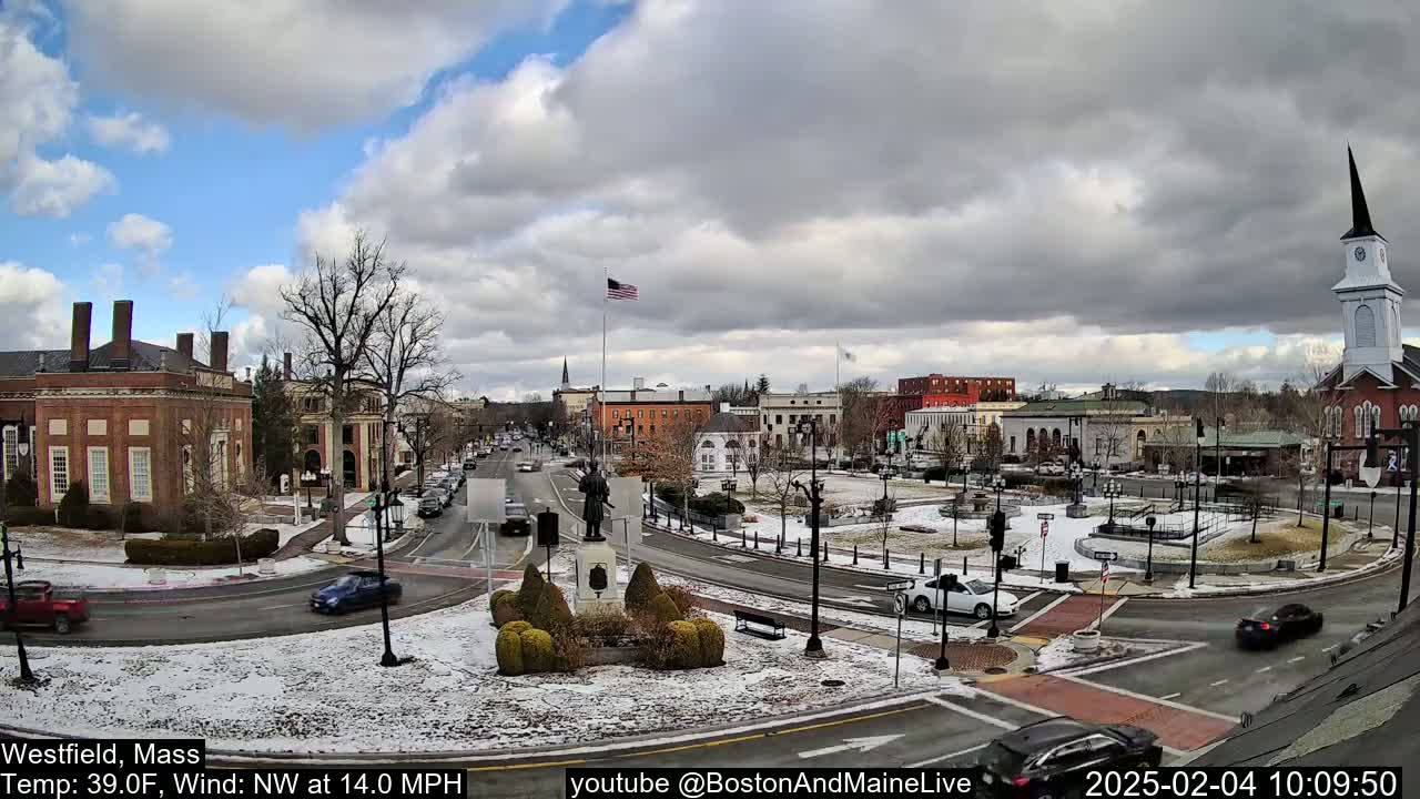 A partly cloudy, cold day in a town square with snow on the ground and cars driving around a central monument, with several brick buildings and a church steeple visible in the background.