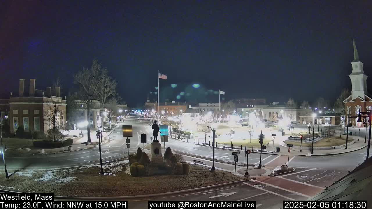 A nighttime view of a town square with a statue in the center, under a clear, dark sky with a temperature of 23°F and a north-northwest wind of 15 mph.