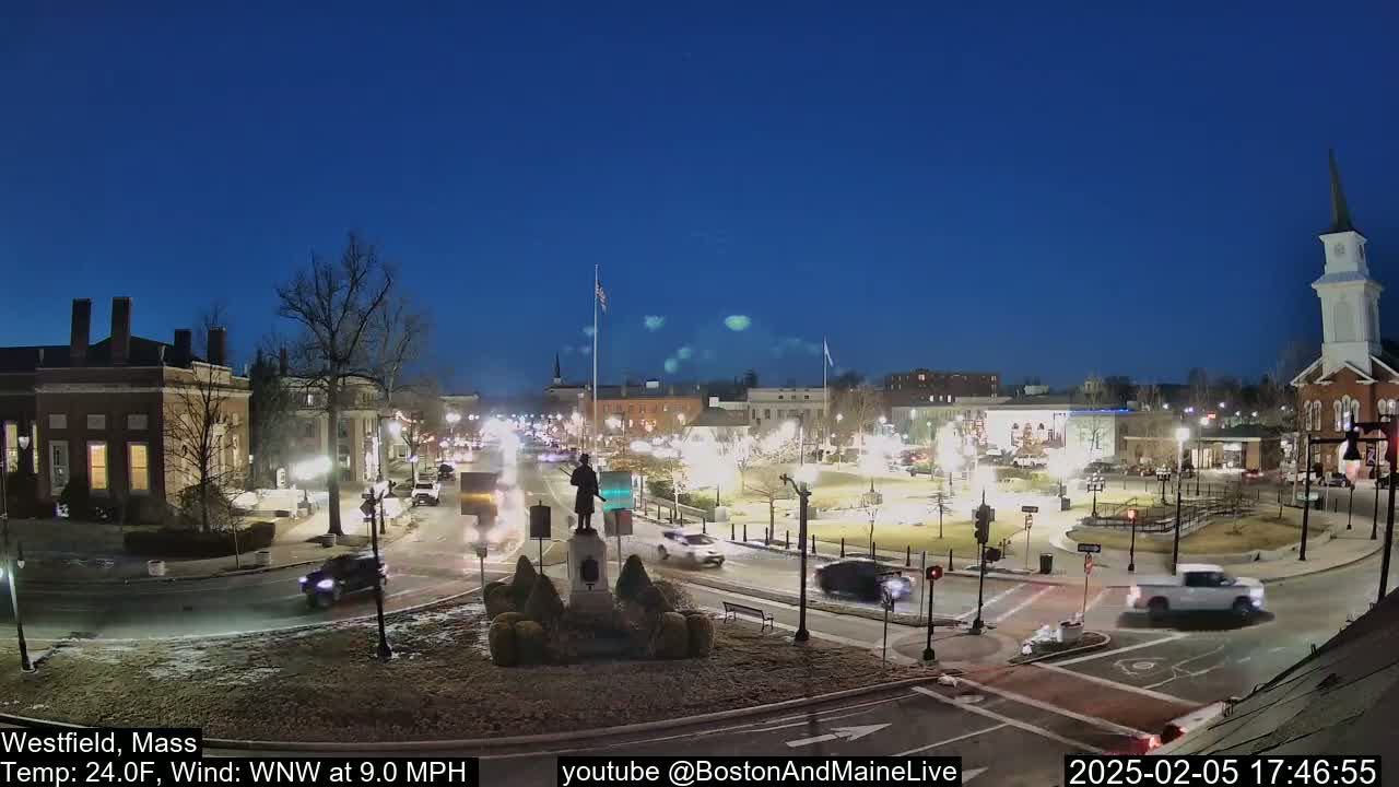 A nighttime view of a town square with light traffic, a statue in the center, and buildings illuminated under a clear, twilight sky.