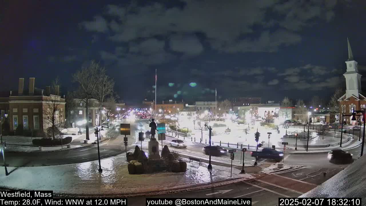 A nighttime view of a snow-covered town square with cars driving around a monument, under a partly cloudy sky.