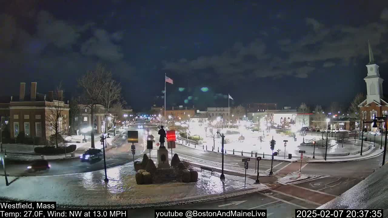 A nighttime view of a snow-covered town square with a statue in the center, under a partly cloudy sky, with a 27°F temperature and a northwest wind at 13 mph.