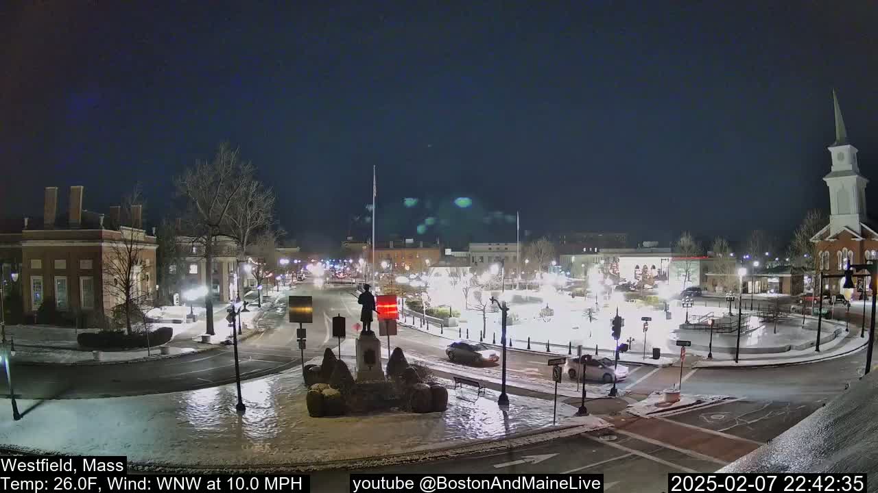 A snow-covered town square at night, illuminated by streetlights, features a statue in the center, with cars driving around it, under a clear, dark sky.