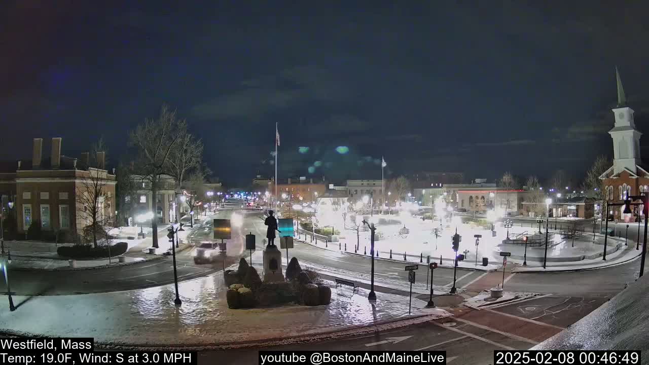 A snow-covered town square at night, with a statue in the center, is lightly trafficked by cars under a dark, clear sky.