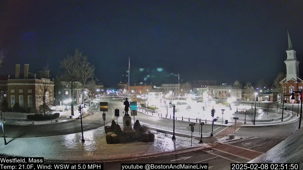 A snow-covered town square at night, illuminated by streetlights, with a statue in the center and buildings surrounding it, under a clear, dark sky.