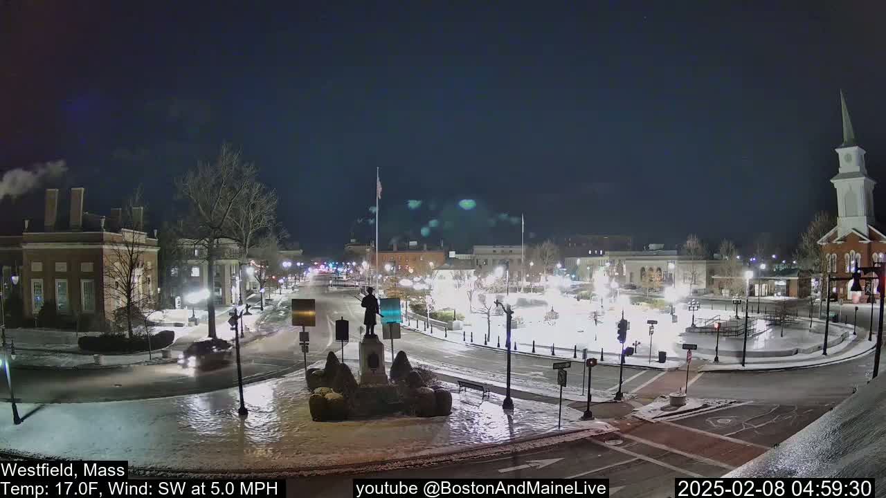 A snow-covered town square at night, illuminated by streetlights, with a statue in the center and a church steeple in the background, under a clear, dark sky.