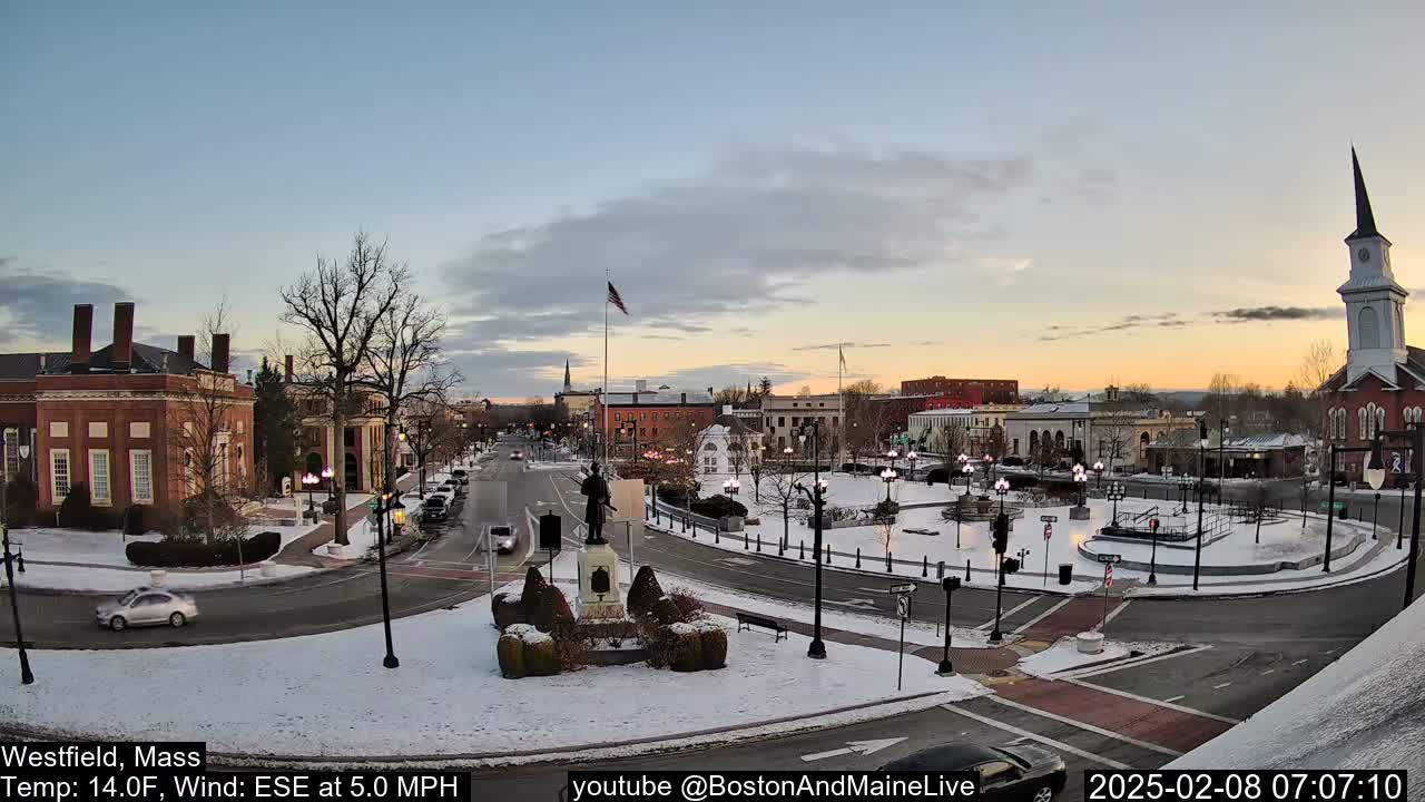 A snow-covered town square with a statue in the center, surrounded by brick buildings, under a partly cloudy twilight sky.