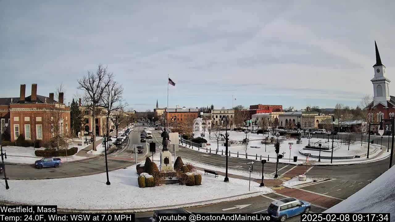 A snow-covered town square with brick buildings, a monument, and cars is seen on a partly cloudy day.