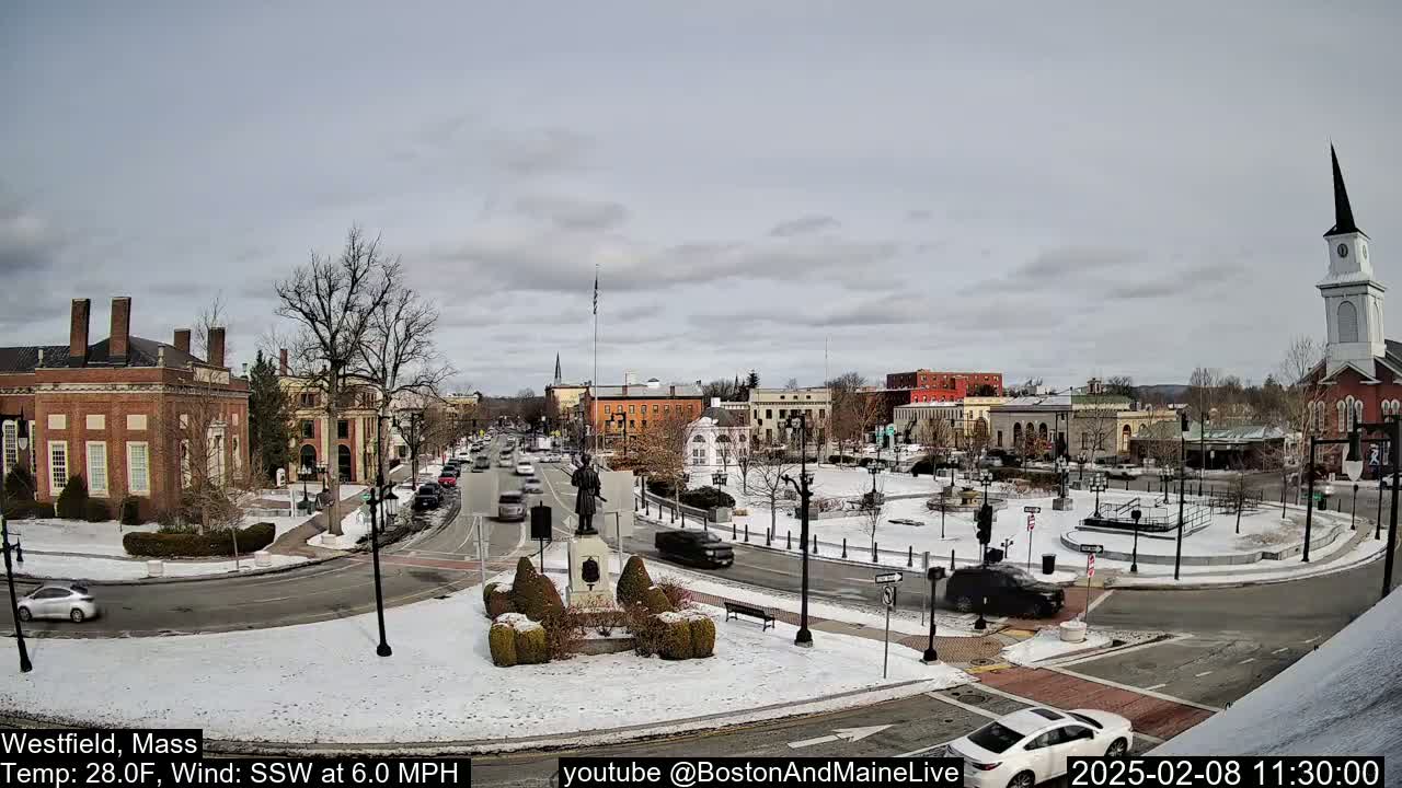 A snow-covered town square with brick buildings, a monument, and cars driving around a traffic circle under an overcast sky.