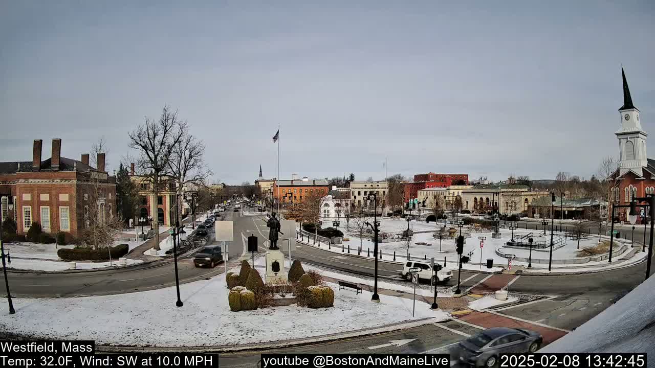 A snow-covered town square with several brick buildings, a statue, and cars driving on roads around it, under a partly cloudy sky.