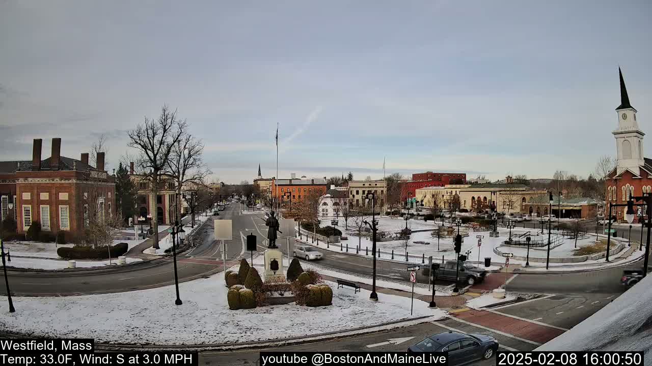 A snow-covered town square with brick buildings, a monument, and a church steeple under a cloudy sky.