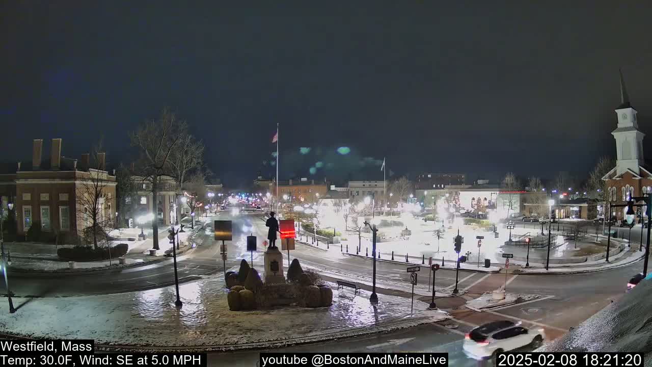 A snow-covered town square at night, with a statue in the center, illuminated by streetlights under a dark sky.