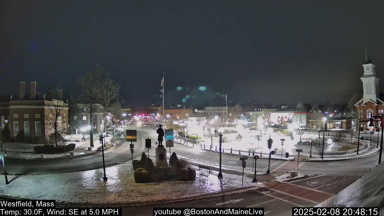 A snow-covered town square at night, illuminated by streetlights, with a statue in the center and light traffic.