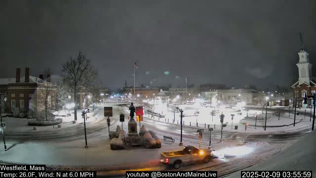 A snow-covered town square at night, with a pickup truck plowing snow and a statue in the center, under a light snowfall with a north wind.