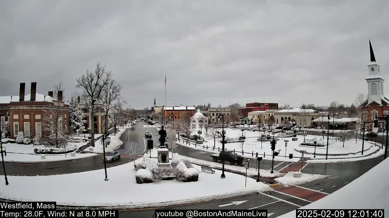 A snow-covered town square with brick buildings, a statue, and a church steeple under a cloudy sky.