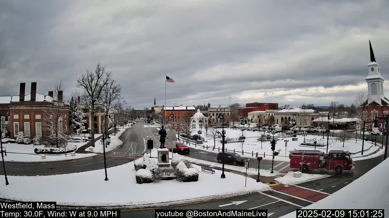 A snow-covered town square with buildings, vehicles, and a monument, under a cloudy sky.
