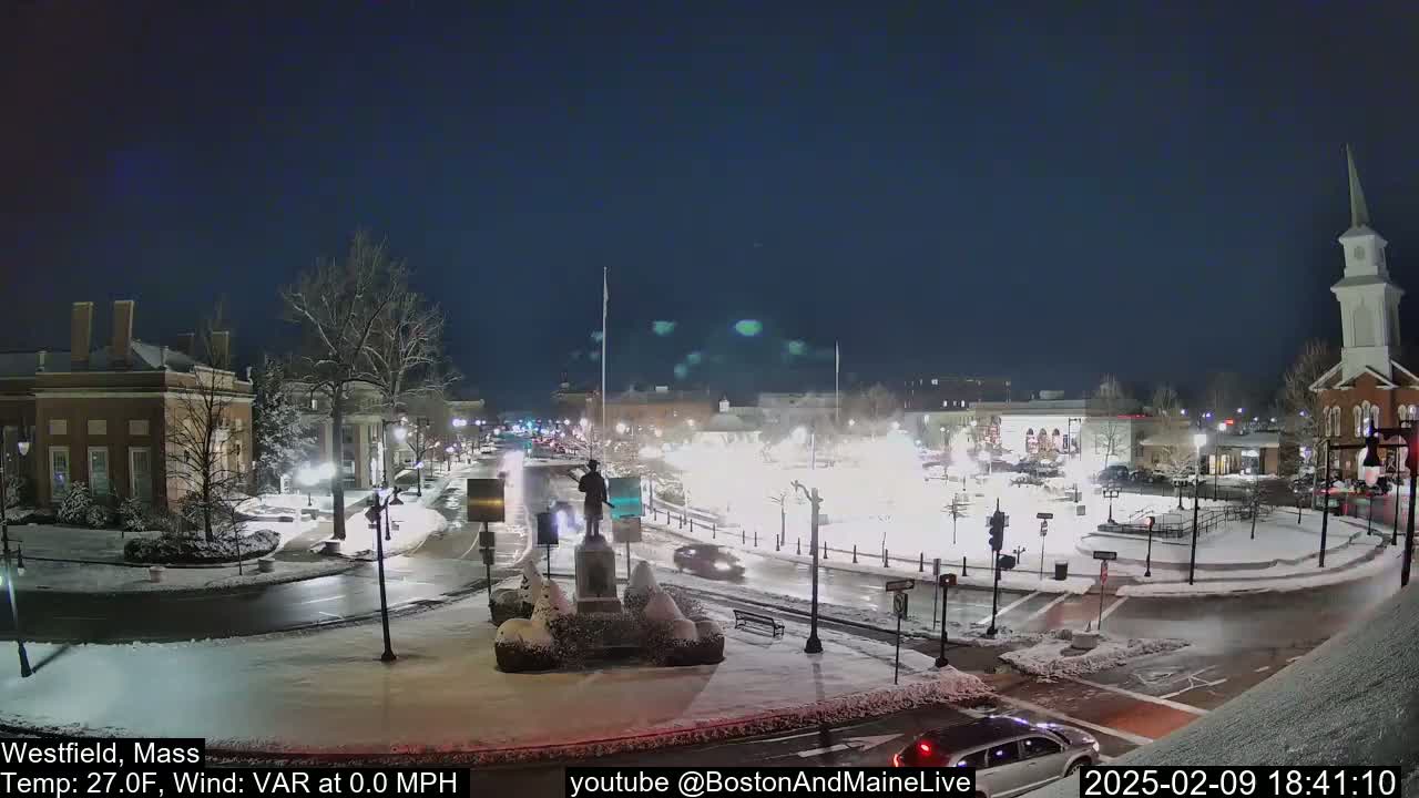 A snow-covered town square at night, illuminated by streetlights, features a statue at its center and several vehicles driving around the roundabout.