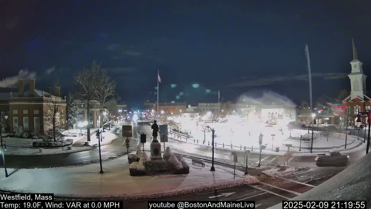 A snow-covered town square at night, illuminated by streetlights, with a statue in the center and a few vehicles on the roads.