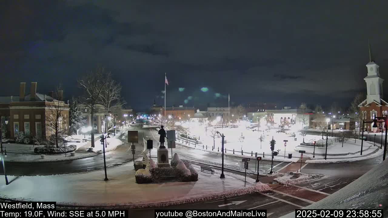 A snow-covered town square at night, illuminated by streetlights, features a statue in the center and several buildings surrounding it, with a calm wind blowing from the southeast.