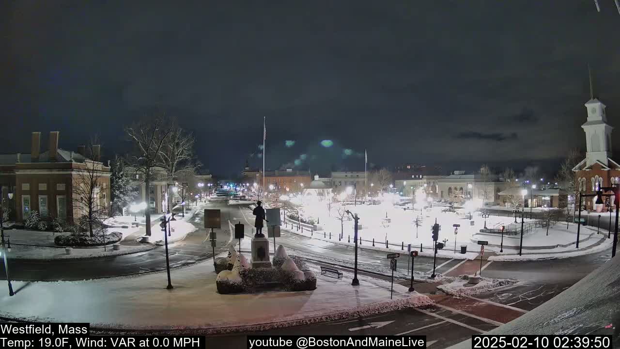 A snow-covered town square at night, illuminated by streetlights, features a statue in the center and buildings surrounding the perimeter.