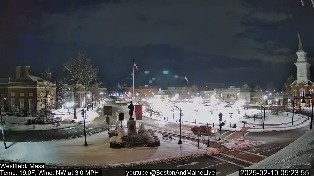 A snow-covered town square at night, illuminated by streetlights, with a statue in the center and a few vehicles on the roads.