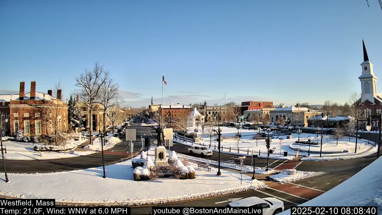A snow-covered town square with brick buildings, a monument, and a church under a clear blue sky.