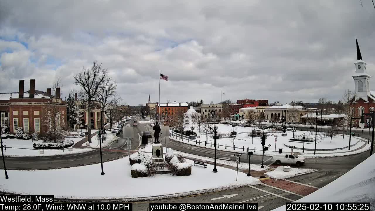 A snow-covered town square with brick buildings, a monument, and a church steeple under a cloudy sky.