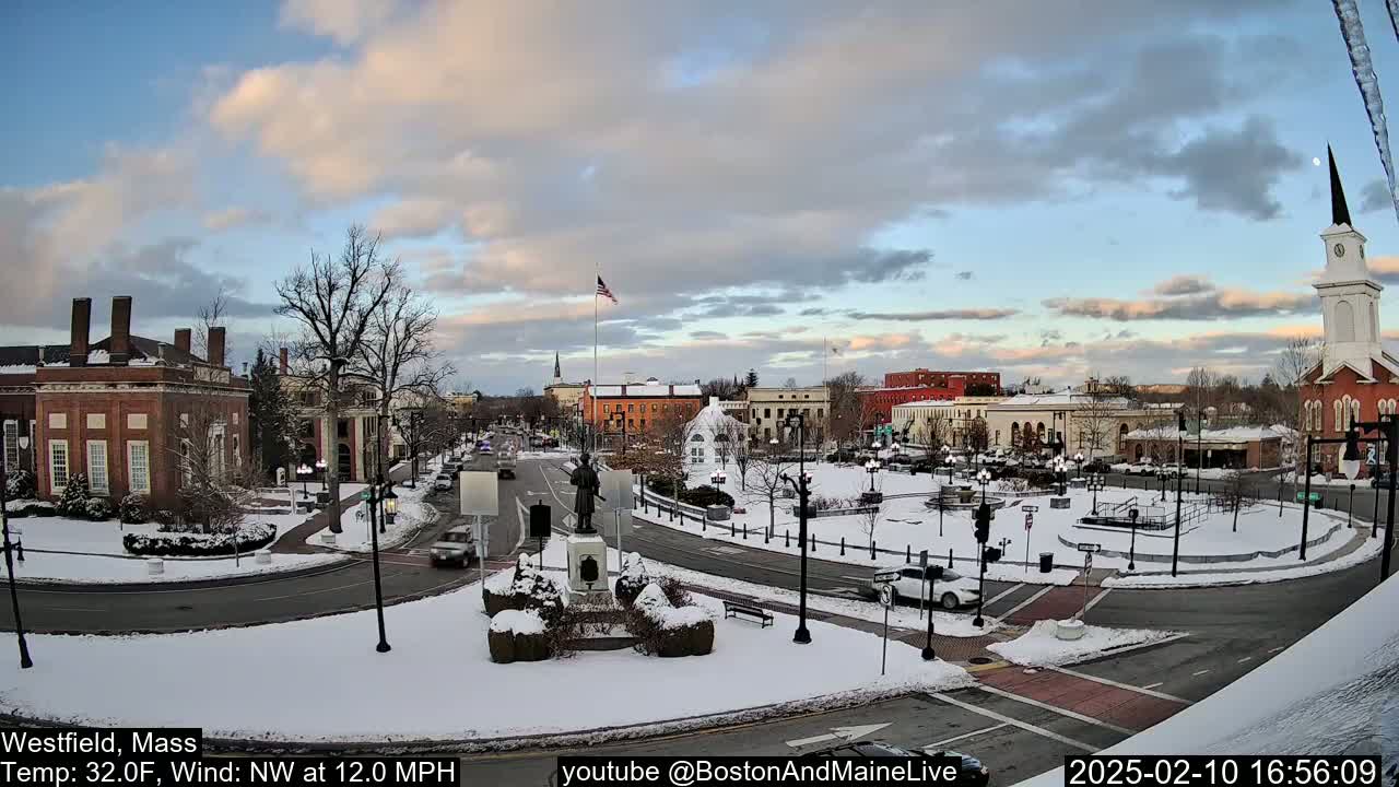 A snow-covered town square with brick buildings, a statue, and a church under a partly cloudy sky, with a northwest wind of 12 mph and a temperature of 32 degrees Fahrenheit.